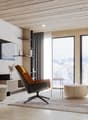 The image shows a modern and cozy living room with a large window overlooking a snowy mountain landscape. The room features a wooden ceiling with recessed lighting, a comfortable-looking leather armchair, and a wooden media console with a mounted TV. The