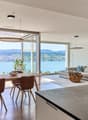 Spacious open-plan living and dining area with large windows offering a scenic view of the lake and surrounding mountains. The dining table with wooden chairs is situated near the windows, while the living room features a comfortable sofa and armchairs. T