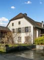 Old house, white walls, brown roof, multiple windows, chimneys, small garden, concrete wall, bushes