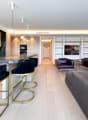modern kitchen with marble countertop, wooden cabinets, 4 bar stools, living room with couch, 2 armchairs, TV, wooden floor