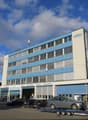 Modern office building with blue and white facade, multiple windows, ground-floor garage, multiple cars parked in front