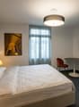 Spacious bedroom with a large bed, leather headboard, and a sitting area with a round table and two chairs. The room has large windows with curtains, and a flat-screen TV on the wall. The decor includes a framed artwork and a drum-shaped light fixture.