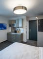 The image shows a modern studio apartment with a kitchenette, a bed, and a living area. The kitchenette has a microwave, a sink, and a dishwasher. The living area has a TV mounted on the wall and a small dining table with chairs. The room has large window