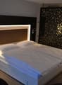Modern bedroom with a large bed, white bedding, and a backlit headboard. The room also features a decorative screen on the wall and a kitchenette area.
