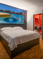 Large bedroom with a king-size bed, hardwood flooring, and a large wall-mounted TV displaying a scenic image of a lake and mountains. The room also has a closet and a bathroom with a red accent wall.
