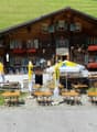 Traditional Swiss chalet-style building with wooden exterior, decorated with flower boxes and antlers. Outdoor seating area with yellow umbrellas and tables. Appears to be a restaurant or cafe in a rural, mountainous setting.