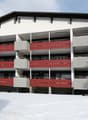 Multi-story building, white exterior, red balconies, covered balconies, located in snowy area, front garden