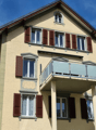 3 story beige house with brown shutters, glass balcony on the second floor, beige pillars, brown roof