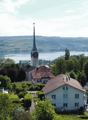 Aerial view of a picturesque town nestled among lush greenery, with a church steeple prominently visible in the center. The town is situated near a large body of water, likely a lake or river, with rolling hills and mountains in the background.