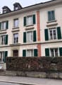 4-storey apartment building, multiple windows with green shutters, central entrance with a small garden, cobblestone street, adjacent buildings