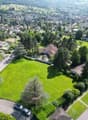 An aerial view of a suburban neighborhood with houses, green fields, and trees. The houses are mostly detached single-family homes with well-kept yards. The area is surrounded by trees, and there are hills in the background. A driveway and car are visible