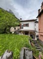 Two-story house with white walls, red roof tiles, and wooden shutters. The house is surrounded by lush greenery, including trees and plants. There is a small balcony or terrace visible on the upper level. The house is situated in a mountainous, rural area