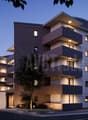 Multi-story apartment building with modern, angular design featuring balconies and terraces, outdoor lighting, and a parking area in the foreground