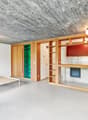 Open-concept studio apartment with concrete ceiling, wooden built-in shelving and cabinets, a bed frame, and a white chair. The room has a minimalist and modern design aesthetic.