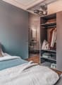 Bedroom with double bed, open door, wooden floor, wardrobe with clothes, bedside table with potted plant, and open bathroom with toilet.