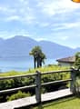 The image shows a scenic outdoor landscape with a view of a lake surrounded by mountains. There is a wooden railing or fence in the foreground, with lush greenery and palm trees in the middle ground. The background features a clear blue sky with some clou