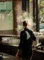 The image shows the interior of a restaurant kitchen. A chef in a black uniform is standing in front of a large commercial kitchen appliance, with steam or smoke visible. The kitchen appears to be well-equipped with various cooking utensils, dishes, and g