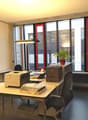 The image shows an office space with a desk, chair, printer, and shelving units. The room has large windows with red frames, providing natural light. There is a potted plant on the desk, adding a touch of greenery to the space.