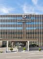 Modern office building with glass facade, multiple floors, entrance with columns, landscaped entrance area with plants and shrubs