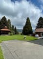 House with parking place, green lawn, small shed, driveway, trees, and mountains in the background
