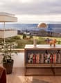 Spacious living room with large windows overlooking a scenic outdoor landscape, including a balcony or terrace. The room features a wooden bookshelf filled with books, a potted plant, and a decorative figurine on a wooden console table.
