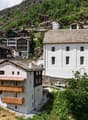 The image shows a picturesque alpine village nestled in a mountainous landscape. The buildings have traditional Swiss architectural styles, with wooden balconies and sloped roofs. There are several multi-story buildings, including a large white building w