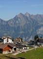 The image shows a picturesque mountain town nestled in a valley surrounded by rugged, snow-capped peaks. The town features a mix of traditional wooden chalets and modern buildings, with a parking area visible in the foreground. The landscape is dominated
