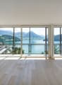 Spacious living room with large windows offering a panoramic view of a lake surrounded by mountains. The room has a minimalist design with light-colored hardwood floors and white walls. The windows provide abundant natural light and a serene, picturesque
