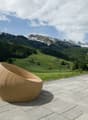 The image shows an outdoor patio area with a wicker chair overlooking a scenic mountain landscape. The patio is made of stone tiles and there is a small potted plant nearby. In the background, there are snow-capped mountains and a forested valley with gre