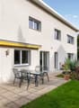 Beige house, large windows, sliding doors, paved patio with table and chairs, awning, garden with plants