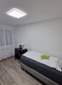 Spacious bedroom with white walls, large windows with sheer curtains, hardwood flooring, a black nightstand, and a bed with white bedding and a green decorative pillow.