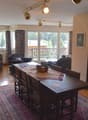 The image shows a spacious open-plan living and dining area. The room has large windows that provide a view of a balcony or terrace overlooking a wooded area. The living room features comfortable sofas and chairs, while the dining area has a wooden table