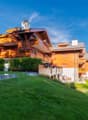 2-story wooden chalet-style house with balconies, surrounded by lush green lawn and trees, set against a blue sky