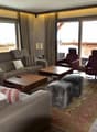 Spacious living room with large windows, hardwood floors, comfortable sofas and armchairs, wooden coffee table, and a balcony or terrace visible through the windows.