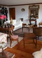 Spacious living room with a mix of traditional and vintage furniture, including a floral patterned sofa, wooden chairs, a coffee table, and a bookshelf. The room features hardwood floors, white walls, and decorative elements such as a large mirror, artwor