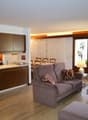 modern kitchen with wooden cabinets, dining area with chairs, two couches, coffee table with books, lamp, large windows