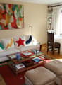 living room: white couch with colorful pillows, glass coffee table, wooden floor, abstract painting on wall, large window, balcony with wooden railing, view of mountains