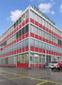 Large office building with red and gray exterior, multiple windows, several cars parked in front