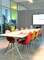 Empty meeting room with glass walls, long white table, orange and blue chairs, screen, whiteboard