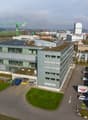 Modern office building with solar panels on the roof, multiple glass windows, several vehicles parked, landscaped area with grass and trees