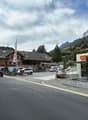 A street with a motorcycle and cars passing by, a building with a flag, a mountain in the background, and a shop with the name 'COOP'.