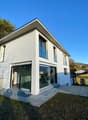 Modern white house, large glass windows, modern architecture, sloping ground with vegetation, small porch with glass doors, concrete fence, mountains in the background.