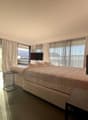 Modern and spacious bedroom with a king-size bed, large windows with white curtains, two armchairs, side table with lamp, small table with chairs, and a TV mounted on the wall. A rug is placed near the chairs.
