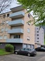 3 story building, beige and gray, balconies, parking lot, cars parked, trees, mountains in the background