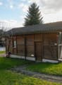 small wooden house, single-storey, slanted roof, windows, wooden entrance, pathway, tree, grass, car parked