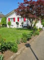 White house with red shutters, driveway, garden with various plants and a tree, neighboring houses in the background