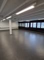 Empty room with white walls, gray floor, white ceiling with beams, large windows, and fluorescent lights