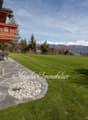 2-story wooden chalet-style house with a large balcony, surrounded by a well-maintained lawn and landscaping. The house has a stone patio in the foreground and offers a scenic view of snow-capped mountains in the background.