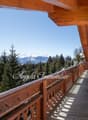 Wooden balcony with a view of snow-capped mountains and a forested landscape. The balcony has a railing with a decorative pattern and there are wooden chairs and furniture on the balcony.