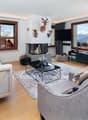 fireplace, 3 deer heads mounted on wall, 3 windows, 2 couches, gray area rug, glass table, gray armchair with fur blanket